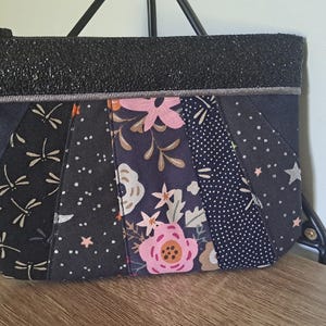 Peut inclure: Une pochette rectangulaire à fermeture éclair avec une bande supérieure noire scintillante et un corps à plusieurs panneaux. Les panneaux présentent divers motifs dont des libellules blanches, des fleurs roses et blanches, des pois et des étoiles.