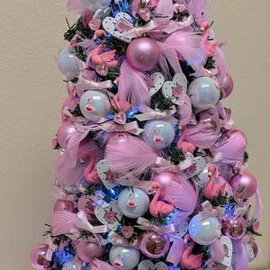 May include: A decorative tabletop tree adorned with pink feathers, iridescent and matte pink ornaments, heart-shaped cutouts, and small flamingo figures. The tree is topped with a flamingo figurine and thin wire stems tipped with white beads.