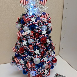 May include: A tabletop artificial Christmas tree decorated with red, white, and blue ornaments, miniature American flags, white star cutouts, and patriotic ribbon bows. A white snowflake topper with integrated LED lights sits at the top.