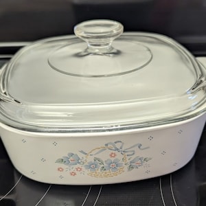 May include: White square baking dish with a glass lid. The dish has a floral design with blue flowers and a basket with a bow. The dish is on a black surface.