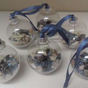 May include: Six clear glass ornaments filled with seashells, sand, and small pieces of greenery. Each ornament has a silver cap and is tied with a blue satin ribbon. The ornaments are arranged on a white surface.