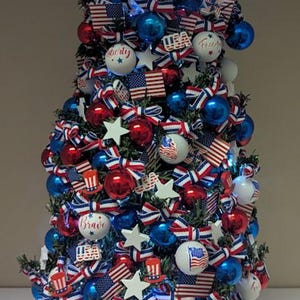 May include: A festive, patriotic tabletop Christmas tree decorated with red, white, and blue ornaments, bows, and miniature American flags. The tree is topped with a white snowflake and sits in a red, white, and blue striped pot.