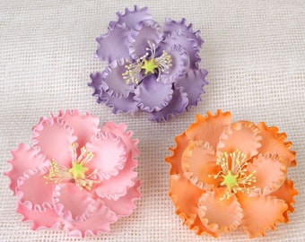 May include: Three fabric flowers in different colors: purple, pink, and orange. The flowers have ruffled petals and are arranged on a white textured background.