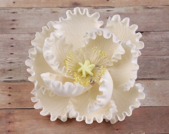 May include: A large, cream-colored flower with ruffled edges and a detailed center. The flower's petals have subtle veining, and the center features yellow stamens and a pale green pistil. The flower is set against a wooden background.