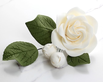 Puede incluir: Una sola rosa blanca con hojas verdes y pequeños capullos blancos. La rosa está hecha de fondant y es una decoración delicada y elegante.