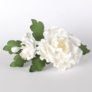 May include: A white sugar flower arrangement with a large peony and smaller white flowers. The flowers are surrounded by green leaves.