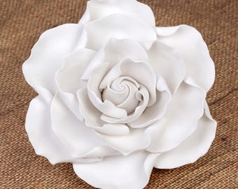 Puede incluir: Una gran rosa blanca de cerámica con múltiples capas de pétalos. La flor está centrada y parece ser un objeto decorativo. Los pétalos tienen una textura suave y están dispuestos en espiral.