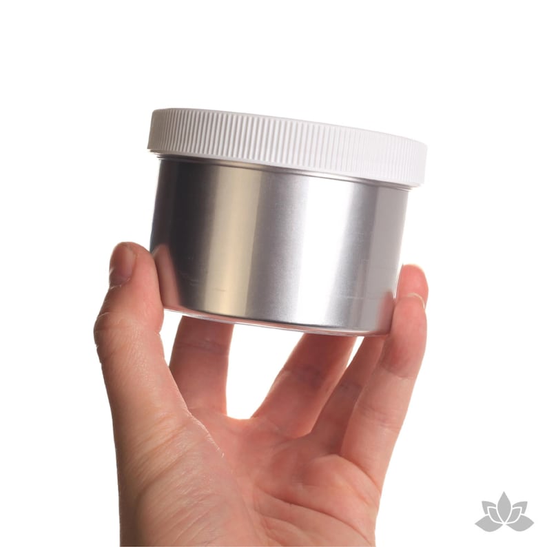 May include: A silver metal container with a white plastic lid. The container is being held in a hand.