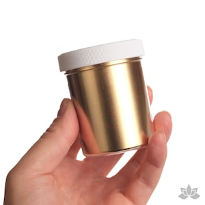 May include: A small jar of gold metallic paint with a white lid. The jar is being held in a hand.