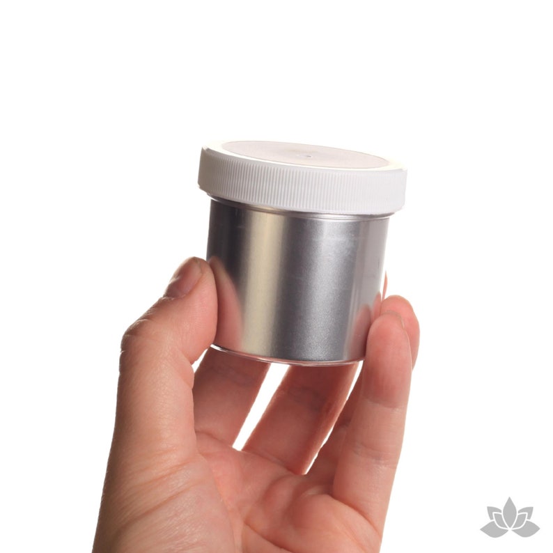May include: A silver metal container with a white lid. The container is being held in a hand.