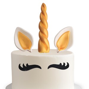 Puede incluir: Un pastel blanco decorado con un cuerno y orejas de unicornio doradas. El pastel también tiene pestañas negras.