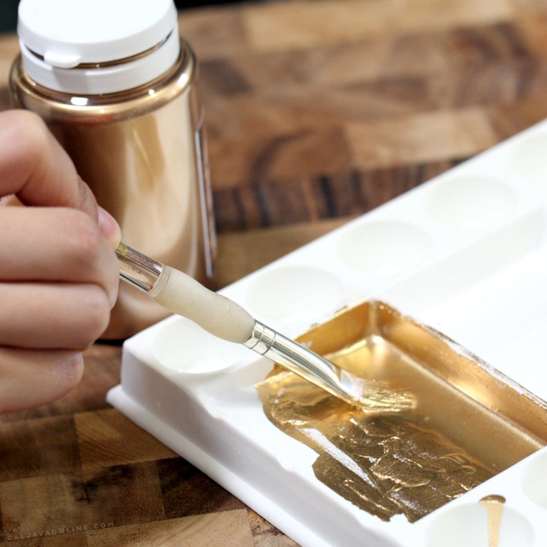 May include: A person using a brush to apply gold paint from a bottle to a white paint palette.