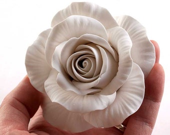 Puede incluir: Una sola rosa artificial blanca, grande y detallada, con un centro en capas. La rosa está hecha de un material liso, similar a la cerámica.
