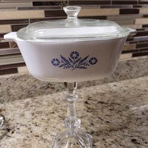 Puede incluir: Plato para hornear blanco con un diseño floral azul y una tapa de vidrio transparente, sentado sobre un pedestal de vidrio transparente.