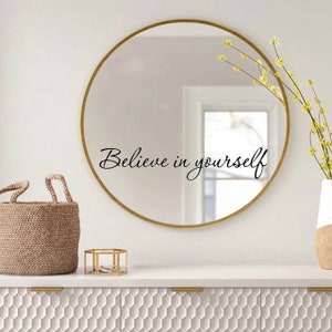 May include: A gold framed round mirror with the words "Believe in yourself" written in black cursive lettering on the mirror surface. The mirror is hanging on a white wall above a white dresser with a hexagonal pattern.