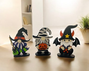 May include: Three wooden Halloween gnome figurines. The gnomes have witch hats, bat wings, and pumpkin details. One gnome holds a broom. The colour scheme includes black, orange, white, purple, and green.