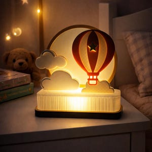May include: A night light shaped like a hot air balloon, with a red and white balloon design and a star. The lamp sits on a white base with a warm glow, and is set against a backdrop of a moon and clouds.
