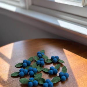 May include: A collection of small, handcrafted felt blueberry clusters, each featuring three round blue felt balls attached to two green felt leaves. These decorative items are arranged in a scattered pattern, showcasing the texture of the wool.