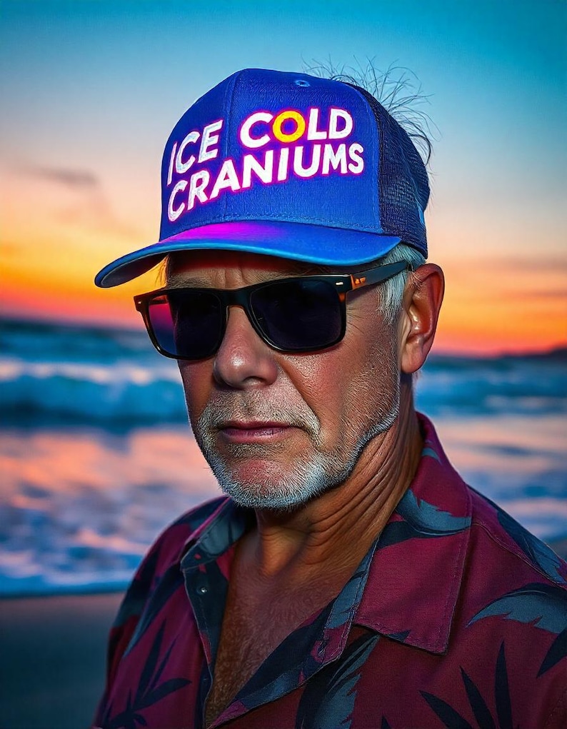 May include: A man wearing a blue and black trucker hat with the text "ICE COLD CRANIUM" in white and yellow letters. He is wearing sunglasses and a red and black patterned shirt. The background is a blurry sunset over the ocean.