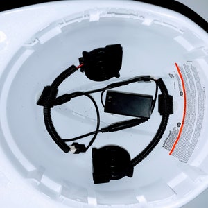 May include: A white hard hat with a black battery pack and two black fan units inside. The fan units are connected to the battery pack with black wires. The hard hat has a warning label on the side.