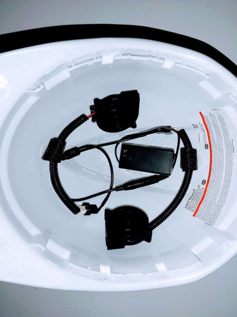 May include: A white hard hat with a black battery pack and two black fan units inside. The fan units are connected to the battery pack with black wires. The hard hat has a warning label on the side.