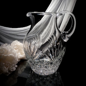 May include: A clear crystal pitcher with a cut glass design. The pitcher has a wide base and a narrow neck with a handle.