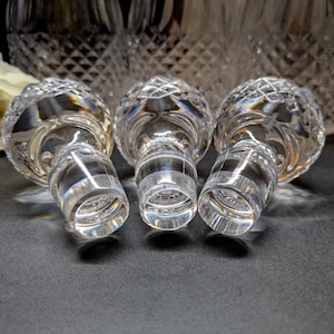 May include: Three clear crystal bottle stoppers with a diamond-cut design. The stoppers are on a dark surface, with a blurred background of a crystal decanter and a white flower.
