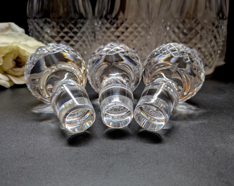 May include: Three clear crystal bottle stoppers with a diamond-cut design. The stoppers are on a dark surface, with a blurred background of a crystal decanter and a white flower.