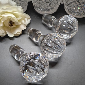 May include: Clear crystal bottle stoppers with diamond-cut patterns. The stoppers have a round, faceted design and a cylindrical stem. Several crystal decanters are visible in the background, along with a white rose.