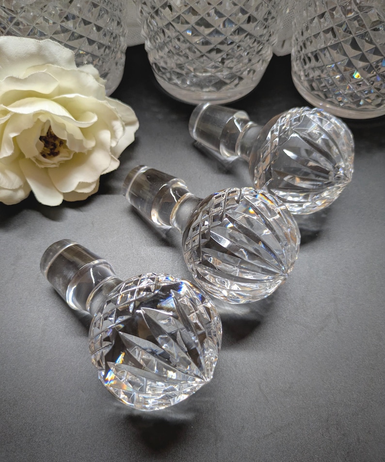 May include: Clear crystal bottle stoppers with diamond-cut patterns. The stoppers have a round, faceted design and a cylindrical stem. Several crystal decanters are visible in the background, along with a white rose.