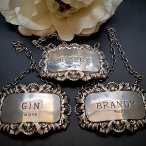 May include: Three antique silver liquor labels with decorative frames and chain fixings. The labels are inscribed with "SHERRY", "GIN", and "BRANDY". The labels are set against a dark backdrop, with a white flower in the background.