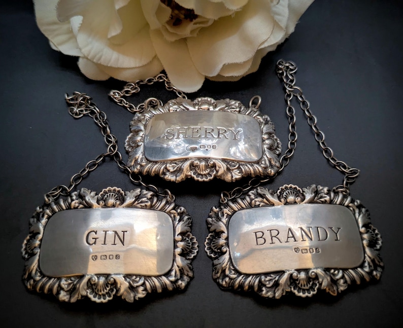 May include: Three antique silver liquor labels with decorative frames and chain fixings. The labels are inscribed with "SHERRY", "GIN", and "BRANDY". The labels are set against a dark backdrop, with a white flower in the background.
