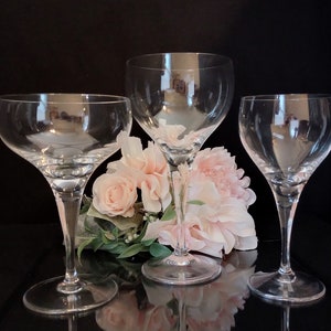 May include: Three clear glass champagne flutes with a delicate stem and a wide bowl. The flutes are arranged in a triangular formation on a black surface with pink flowers in the background.