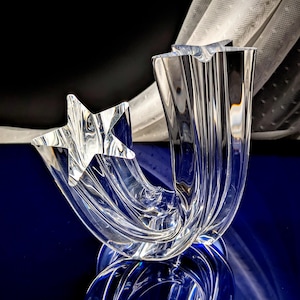 May include: A clear, star-shaped glass sculpture with a curved, flowing design. The sculpture features a five-pointed star and a vertical, fluted element. The piece is set against a dark background with a blue surface.