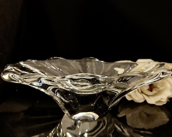 May include: A clear glass bowl with a wavy, organic design. The bowl has a wide, shallow base and a tall, slender stem. The bowl is sitting on a black surface.