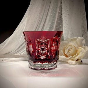 May include: A ruby red, cut crystal tumbler with intricate floral and geometric patterns. The glass has a wide mouth and a faceted base. A white rose and a sheer, white curtain are in the background.