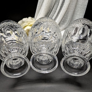 May include: Three clear crystal goblets with detailed cut glass designs. The goblets feature a rounded bowl and a short stem with a wide, circular base. A white flower and sheer fabric are in the background.