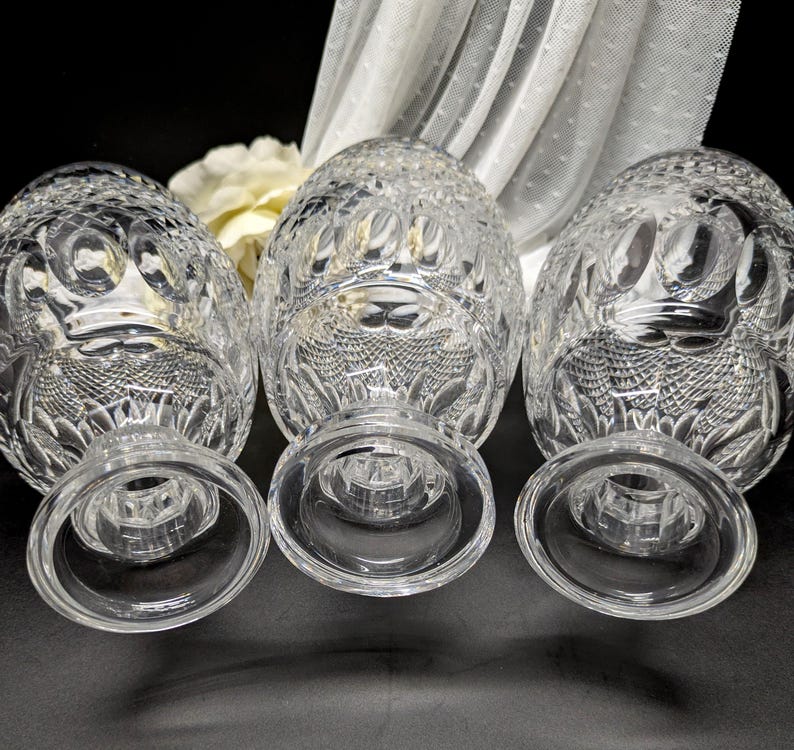 May include: Three clear crystal goblets with detailed cut glass designs. The goblets feature a rounded bowl and a short stem with a wide, circular base. A white flower and sheer fabric are in the background.