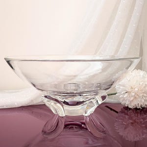 May include: A clear glass bowl with a wide, circular rim and a smooth, transparent surface. The bowl is supported by three distinct, curved glass feet that elevate the base, creating a stable structure for this decorative serving piece.