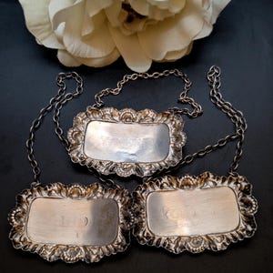 May include: Three antique silver bottle labels with decorative, floral frames and attached chains. The rectangular labels have engraved text, and are set against a dark background with a white flower.