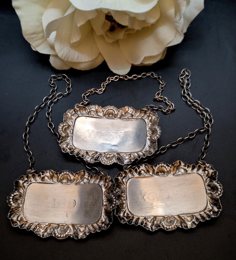 May include: Three antique silver bottle labels with decorative, floral frames and attached chains. The rectangular labels have engraved text, and are set against a dark background with a white flower.