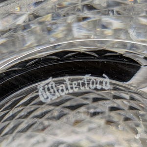 May include: Close-up of a clear crystal glass with a diamond-cut pattern. The word "Waterford" is etched in elegant script on the glass. The glass reflects light, creating a sparkling effect.