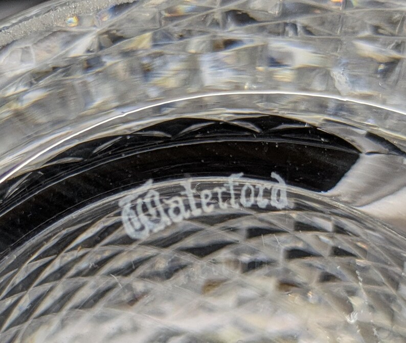 May include: Close-up of a clear crystal glass with a diamond-cut pattern. The word "Waterford" is etched in elegant script on the glass. The glass reflects light, creating a sparkling effect.