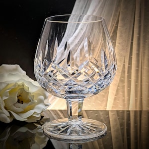 May include: A clear, cut-crystal brandy snifter with a wide bowl and short stem. The glass features a diamond and vertical cut pattern. The base is round and also cut with a starburst design. A white flower and sheer curtain are in the background.