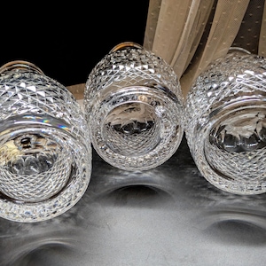 May include: Three clear, cut-glass vases with a diamond pattern. The vases are arranged in a row, with their bottoms facing the viewer. The glass reflects light, creating a sparkling effect. A white flower and sheer fabric are in the background.