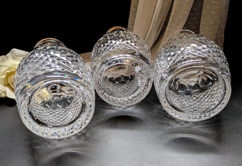 May include: Three clear, cut-glass vases with a diamond pattern. The vases are arranged in a row, with their bottoms facing the viewer. The glass reflects light, creating a sparkling effect. A white flower and sheer fabric are in the background.