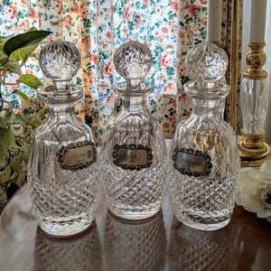 May include: Three clear glass decanters with diamond-cut patterns and crystal stoppers. Each decanter has a silver label that reads "Sherry", "Gin", and "Brandy". The decanters are displayed on a reflective surface.