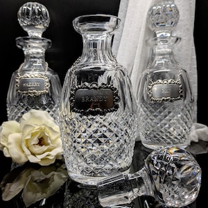 May include: Three crystal liquor decanters with diamond-cut patterns. Each decanter has a silver label that reads "Sherry", "Brandy", and "Gin". One decanter has its stopper removed and lying on its side. A white flower is in the foreground.