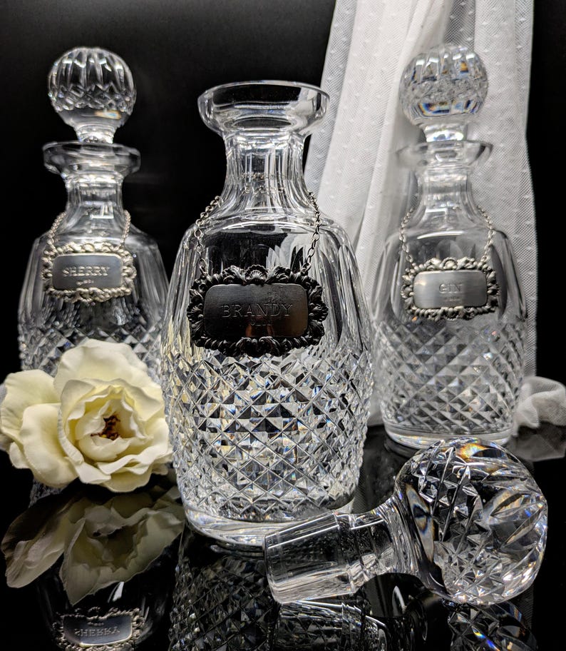 May include: Three crystal liquor decanters with diamond-cut patterns. Each decanter has a silver label that reads "Sherry", "Brandy", and "Gin". One decanter has its stopper removed and lying on its side. A white flower is in the foreground.