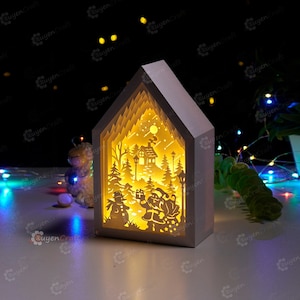 May include: A white wooden light-up house with a cut-out design of a winter scene. The scene includes a snowman, Santa Claus, a house, trees, and a moon. The house is illuminated from within, creating a warm glow.